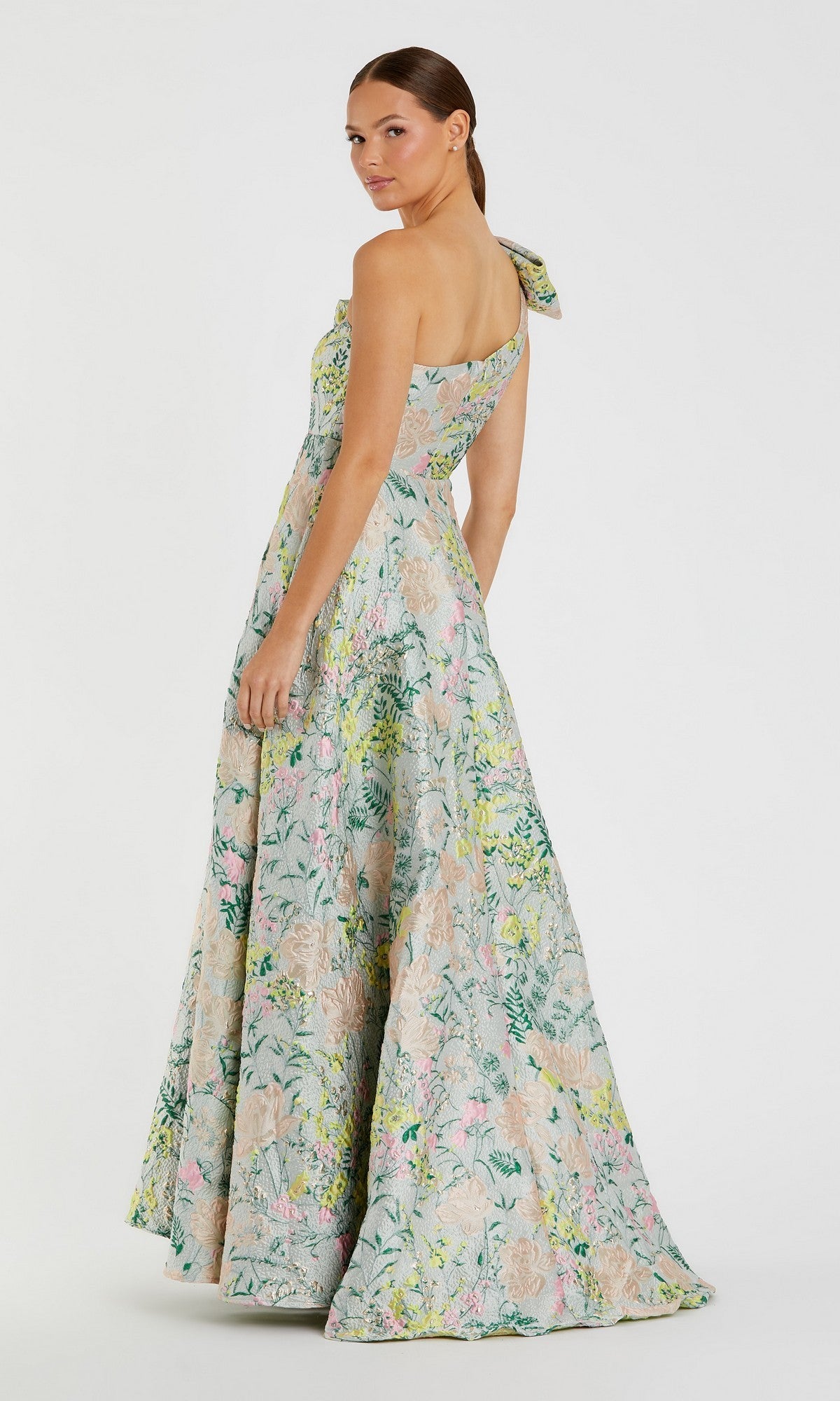 A woman wearing the Mac Duggal 11919 long formal dress, a floor-length one-shoulder gown with a floral brocade design, bow shoulder detail, and elegant A-line skirt, stands against a plain white background.